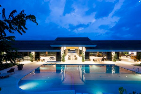 An elegant, well-lit building with a large swimming pool in front, surrounded by tables and chairs under a blue evening sky.