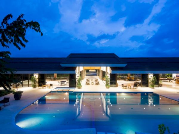 An elegant, well-lit building with a large swimming pool in front, surrounded by tables and chairs under a blue evening sky.
