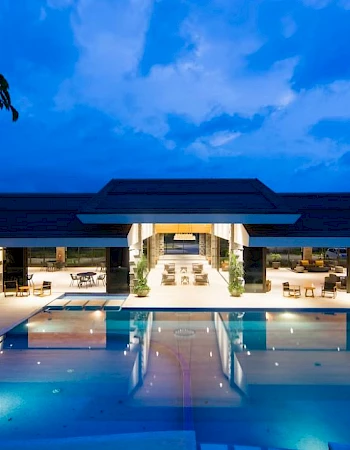 An illuminated luxury resort with an open-air design, centering around a large pool, with seating areas and potted plants under a blue evening sky.