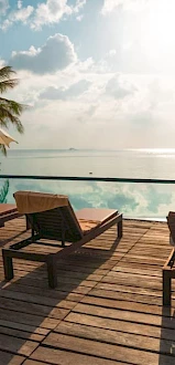 A serene beach scene with lounge chairs and umbrellas on a wooden deck facing a calm, sunlit ocean. Palm trees provide additional shade.