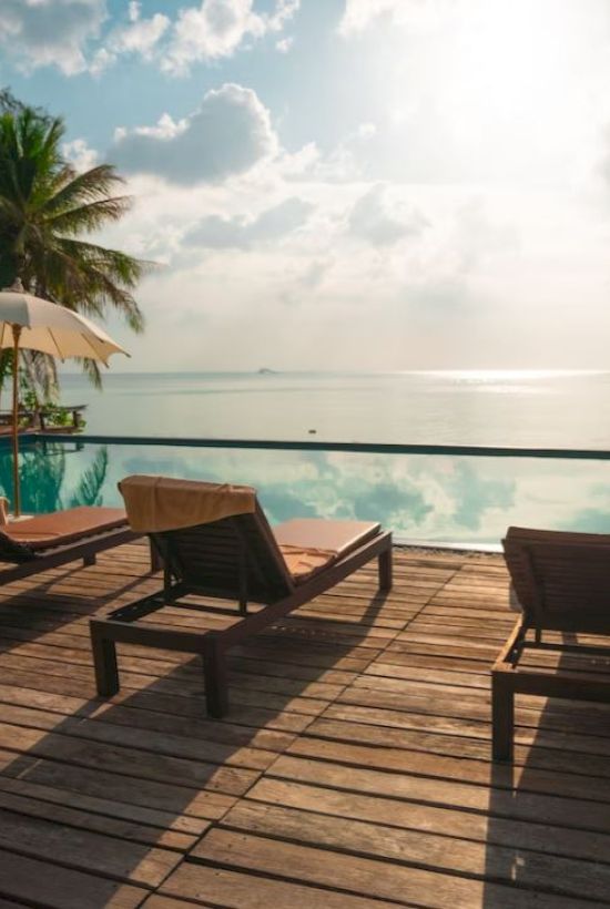 Lounge chairs and umbrellas by a beautiful infinity pool overlooking a serene ocean with a bright sky and scattered clouds.