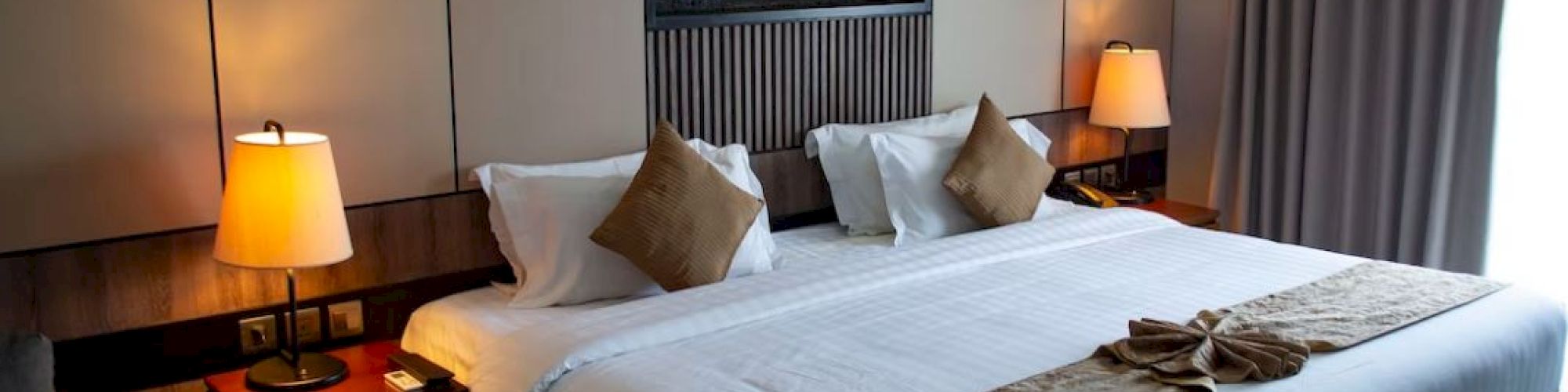 A cozy hotel room with a large bed, two bedside tables with lamps, decorative pillows, and a wall-mounted fireplace, plus a curtain-covered window.