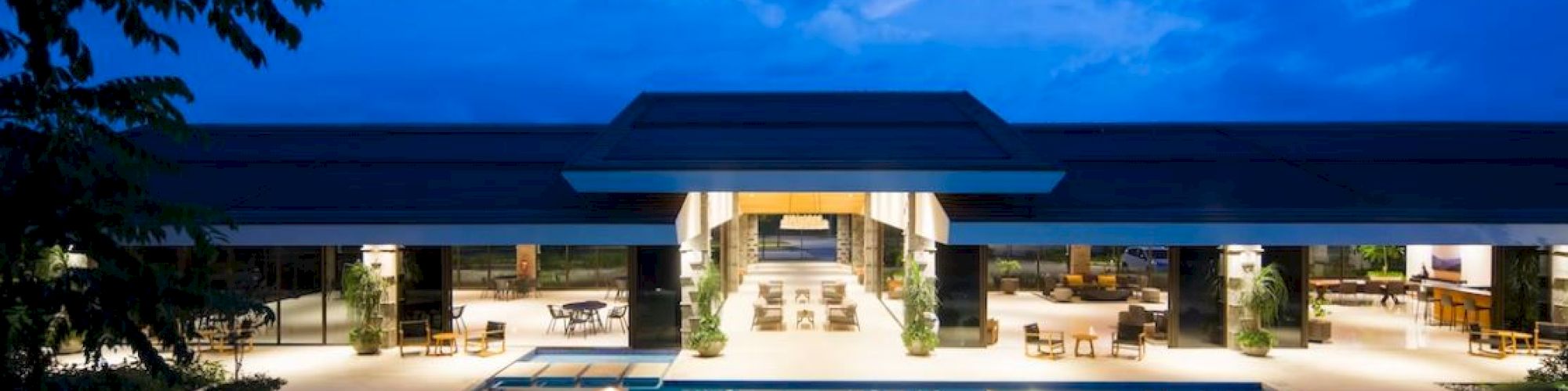 A luxurious building at dusk features a large, illuminated outdoor pool with lounge areas and open-air seating under a modern roof structure.