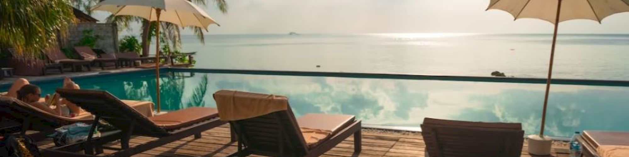 A serene poolside scene with lounge chairs and umbrellas, overlooking a calm sea surrounded by tropical palm trees under a bright sky.
