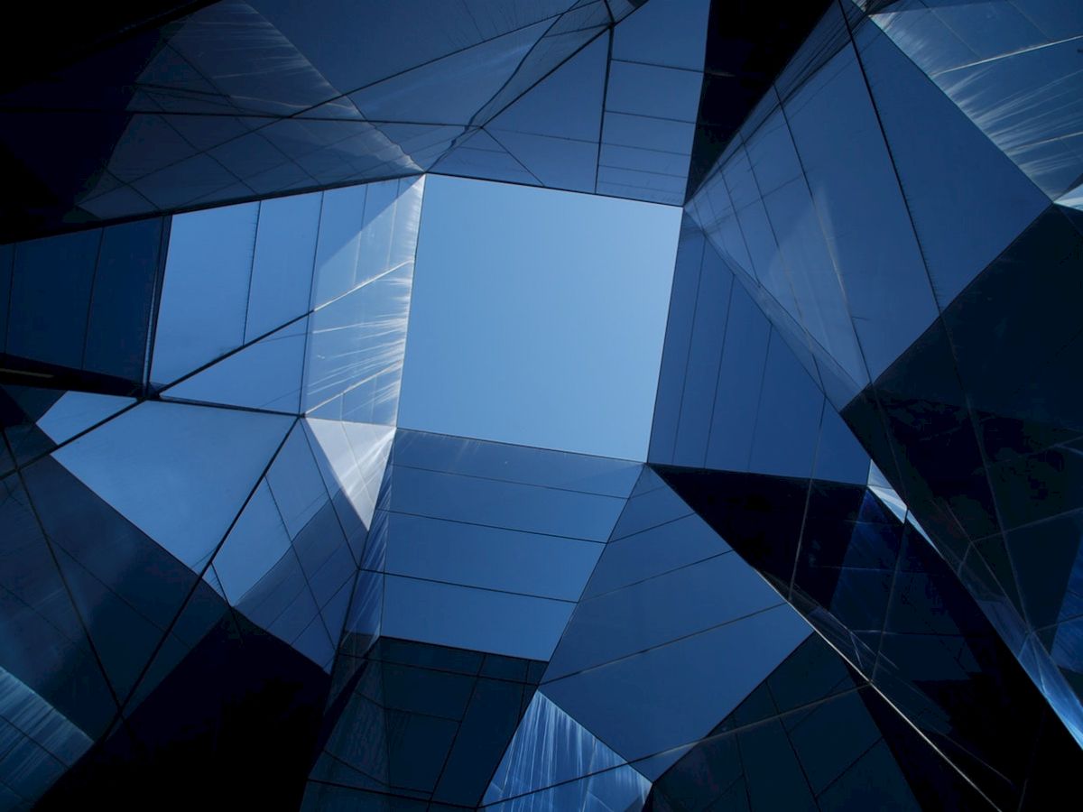 The image shows an upward view of the sky through a geometric arrangement of reflective glass surfaces, creating a modern, abstract architectural effect.