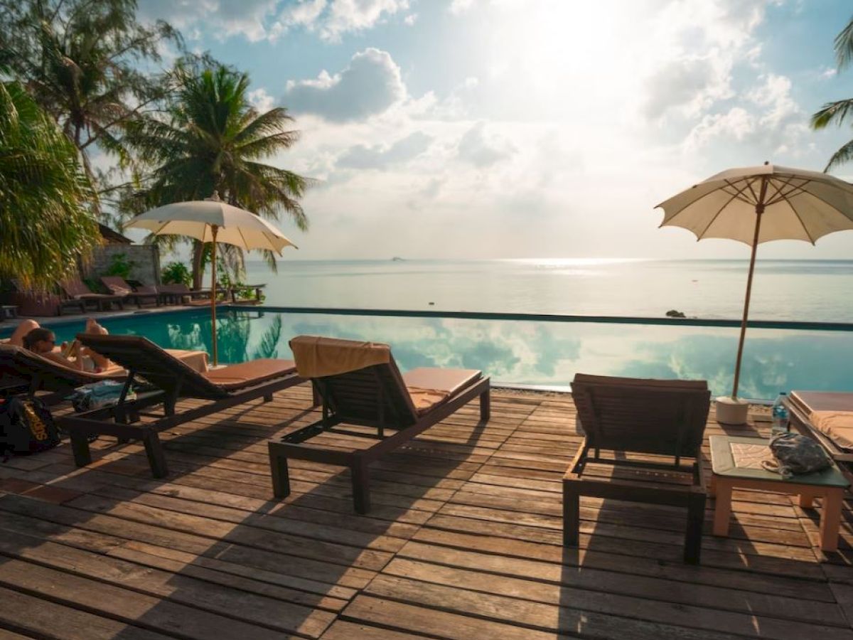 A serene tropical beach scene with lounge chairs, umbrellas, and a swimming pool overlooking a calm ocean under a bright sky.