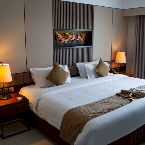 A well-lit hotel room features a large bed with white linens and brown accents, bedside tables with lamps, and a wall-mounted fireplace.