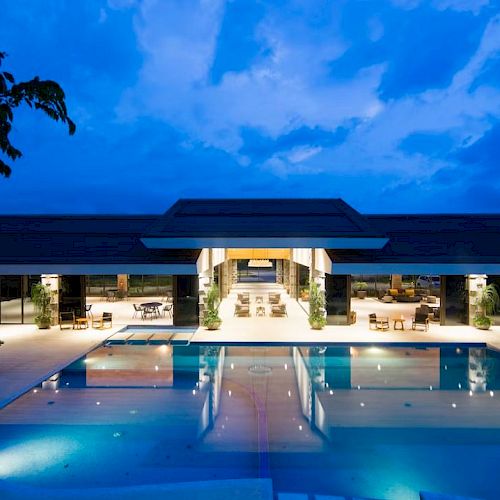 An illuminated modern building with glass walls, featuring a large, rectangular outdoor swimming pool with lounge chairs and tables around it.