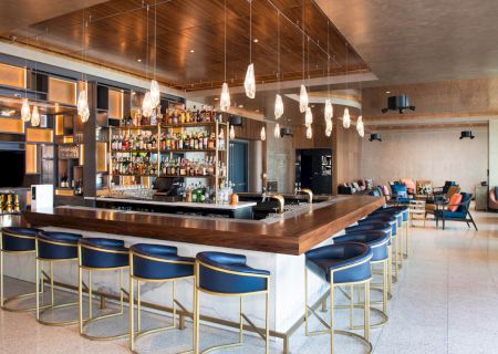 A modern bar with a variety of bottles, blue barstools, pendant lights, and a lounge area with seating in the background.