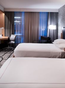 A modern hotel room with two beds, a wall-mounted TV, a desk with a chair, and contemporary lighting and design elements.