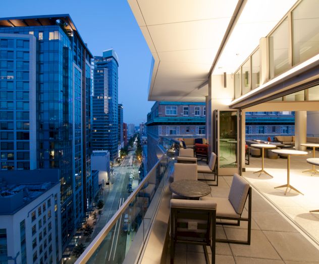 The image shows a modern cityscape at dusk with tall buildings on the left and a sleek, well-lit balcony with chairs and tables on the right.