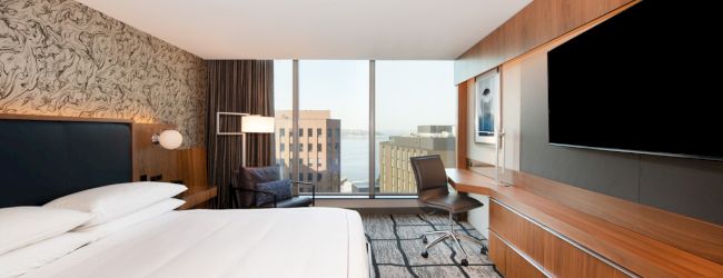 A modern hotel room with a large bed, wall art, a desk and chair, a large TV, and a panoramic window offering a view of city buildings.
