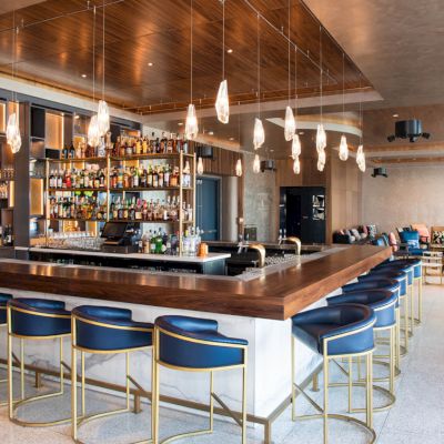 The image shows a modern bar with a counter and chairs, shelves stocked with bottles, pendant lights, and a seating area in the background.