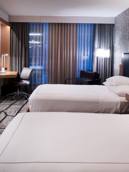 The image shows a modern hotel room with two neatly made beds, a television, a desk with a chair, and contemporary decor and lighting.
