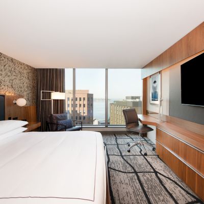 Modern hotel room with a large bed, wall-mounted TV, desk, and a window offering a city view. The decor is contemporary with wooden accents.