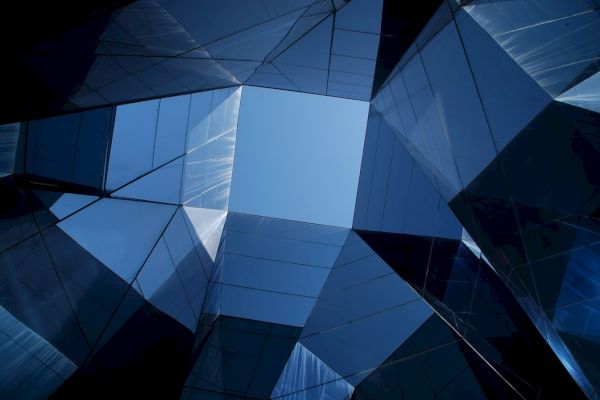 The image shows a geometric pattern formed by glass buildings reflecting each other, with a clear blue sky visible through the center.