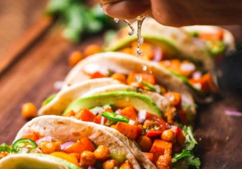 A hand squeezes lime over a row of tacos filled with a colorful mix of vegetables and chickpeas on a wooden board.