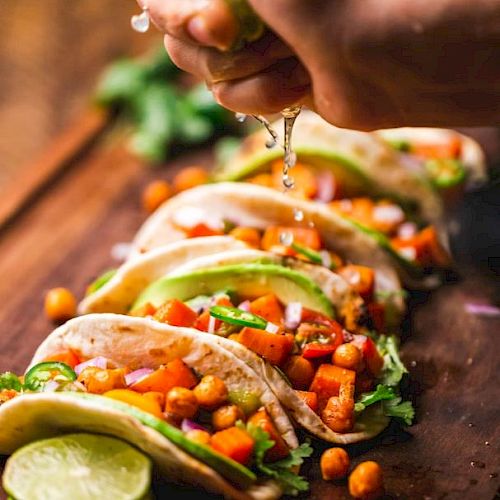 A hand squeezes lime over a row of tacos filled with a colorful mix of vegetables and chickpeas on a wooden board.
