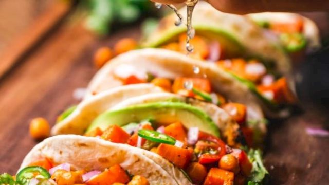 A hand squeezes lime over a row of tacos filled with a colorful mix of vegetables and chickpeas on a wooden board.