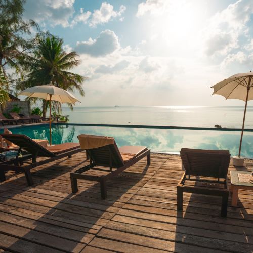Lounge chairs and umbrellas next to an infinity pool overlook a calm ocean with a serene sky and palm trees in the background.