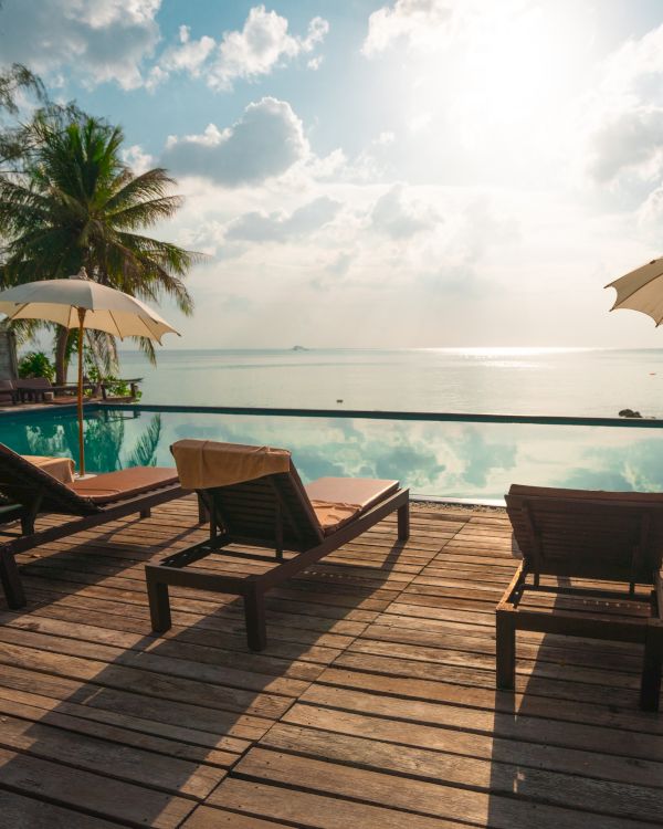Lounge chairs and umbrellas next to an infinity pool overlook a calm ocean with a serene sky and palm trees in the background.