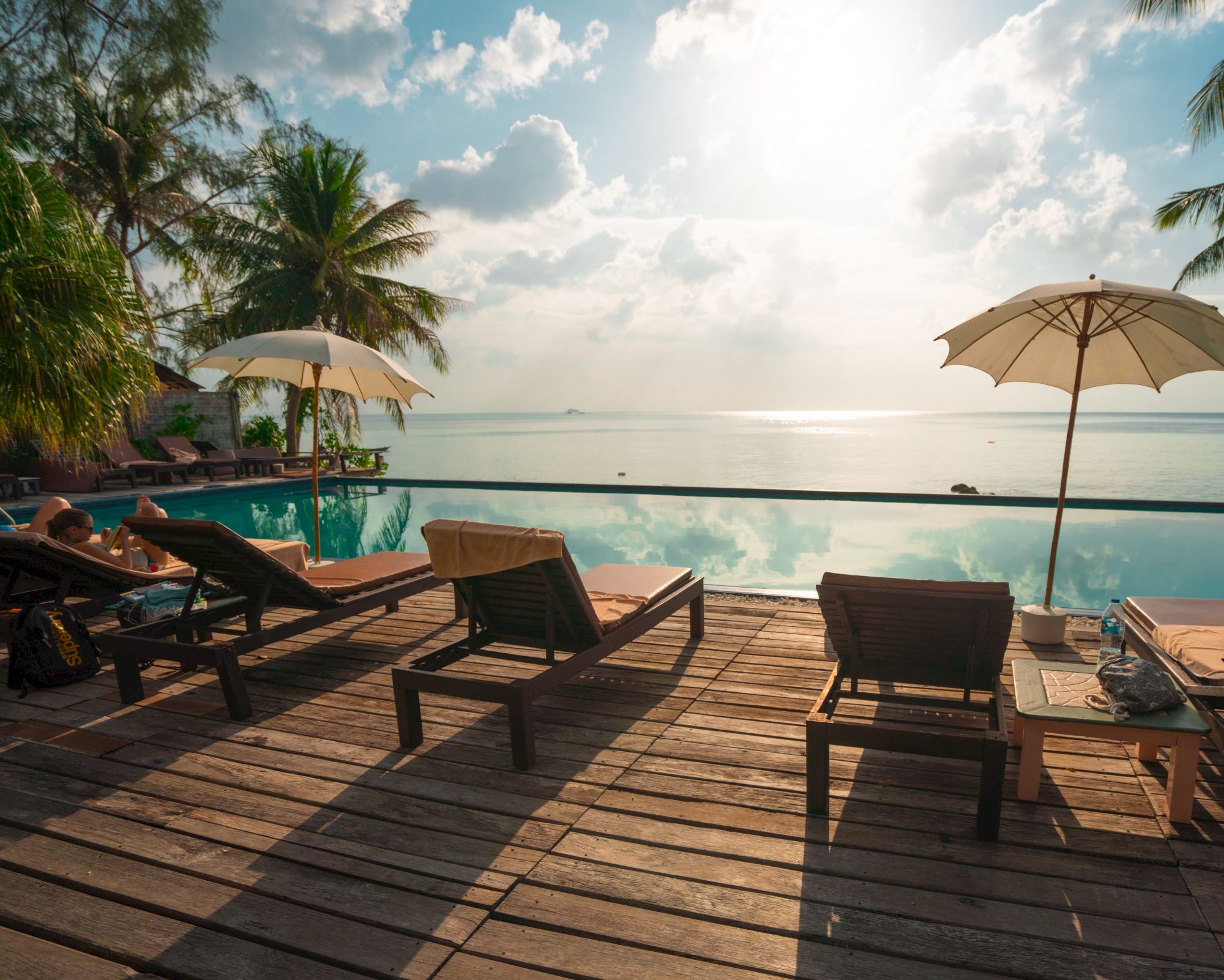 A serene beach scene with lounge chairs and umbrellas on a wooden deck overlooking an infinity pool and the ocean, with palm trees in the background.
