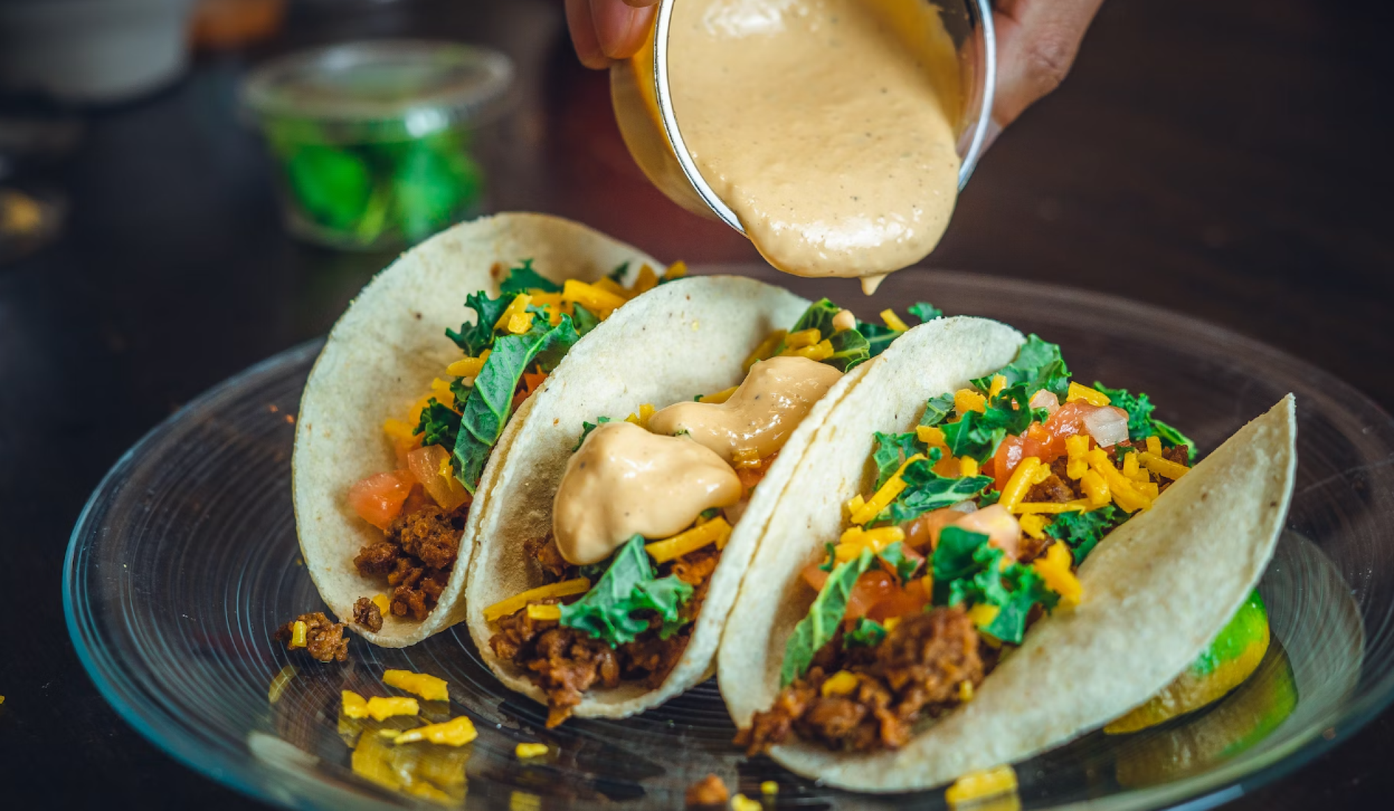 Three soft tacos filled with a mixture of greens, meat, tomatoes, and cheese are being topped with a creamy sauce on a glass plate.