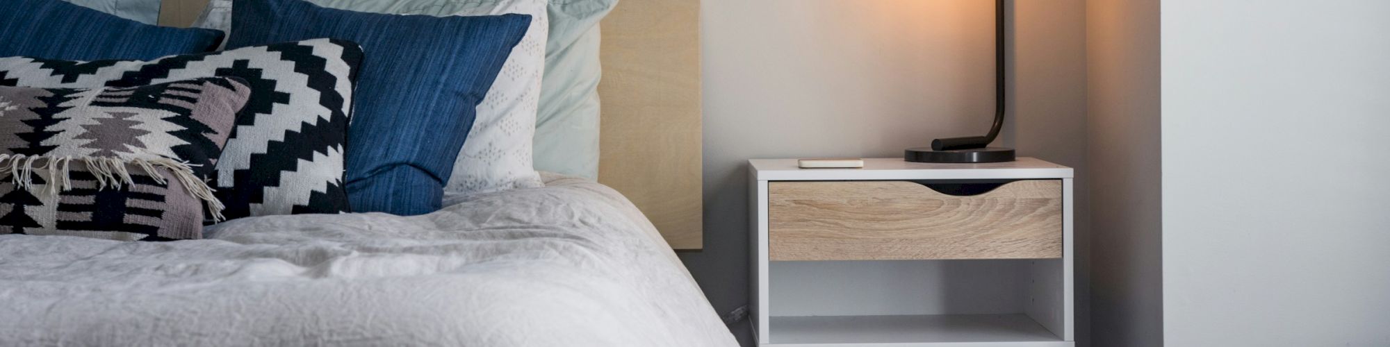 A cozy bedroom with a white bed, blue and patterned pillows, and a wooden nightstand featuring a modern lamp with an exposed bulb.