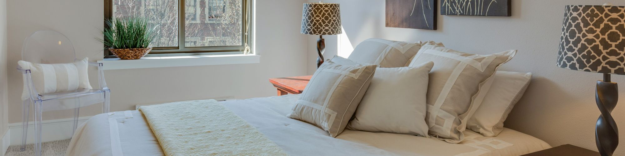 A cozy bedroom with a neatly made bed, decorative pillows, a yellow throw, two art pieces, lamps, a window, a plant, and a chair by the window.