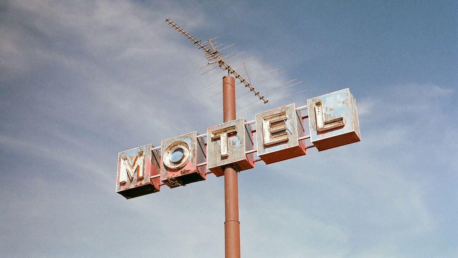 This image shows an old, weathered motel sign mounted on a pole with an antenna on top, set against a partly cloudy sky.