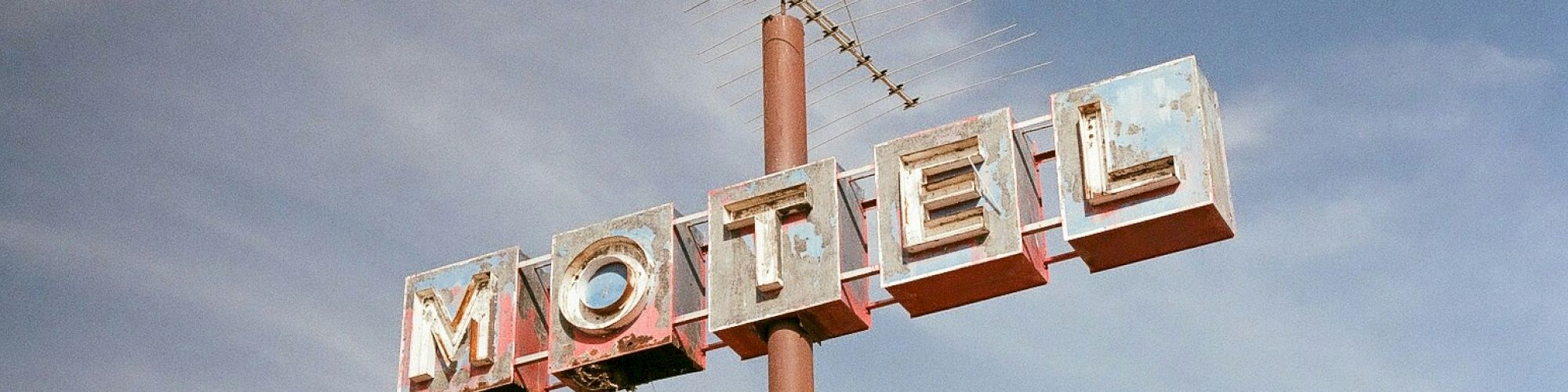 This image shows an old, weathered motel sign mounted on a pole with an antenna on top, set against a partly cloudy sky.