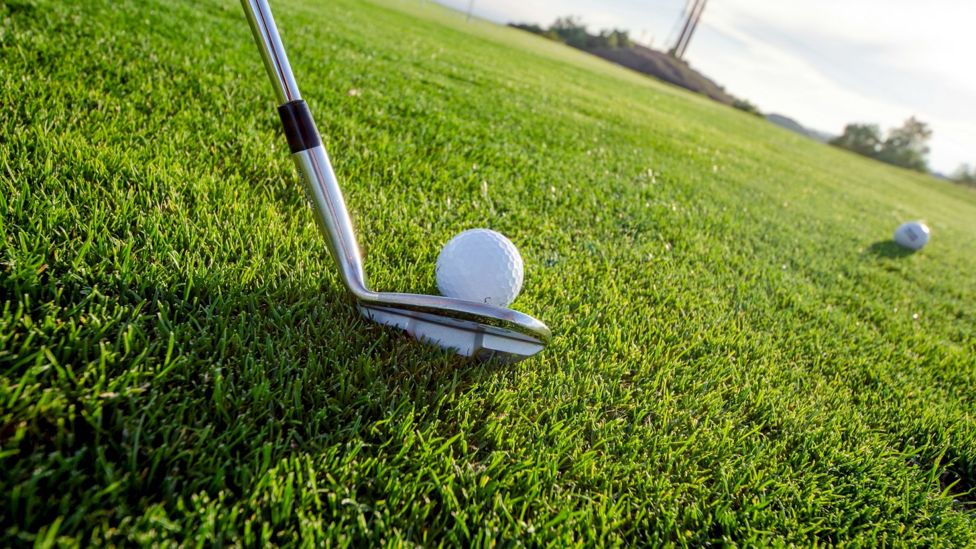 A golf club positioned to hit a golf ball on a lush green course, with another ball in the distance and a clear sky above.
