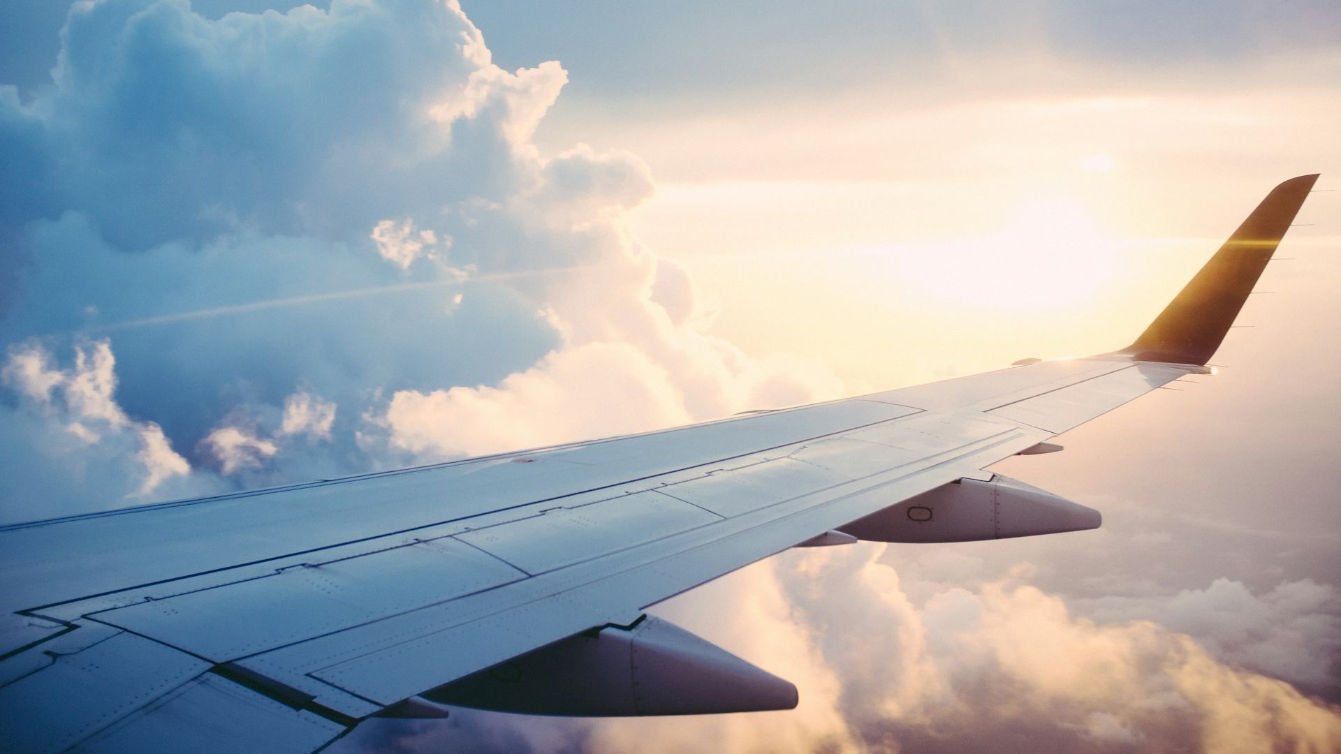 Image shows an airplane wing against a backdrop of fluffy clouds and a bright, warm sunset sky.