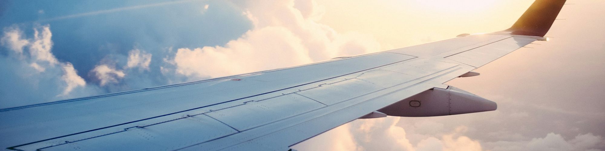 Image shows an airplane wing against a backdrop of fluffy clouds and a bright, warm sunset sky.