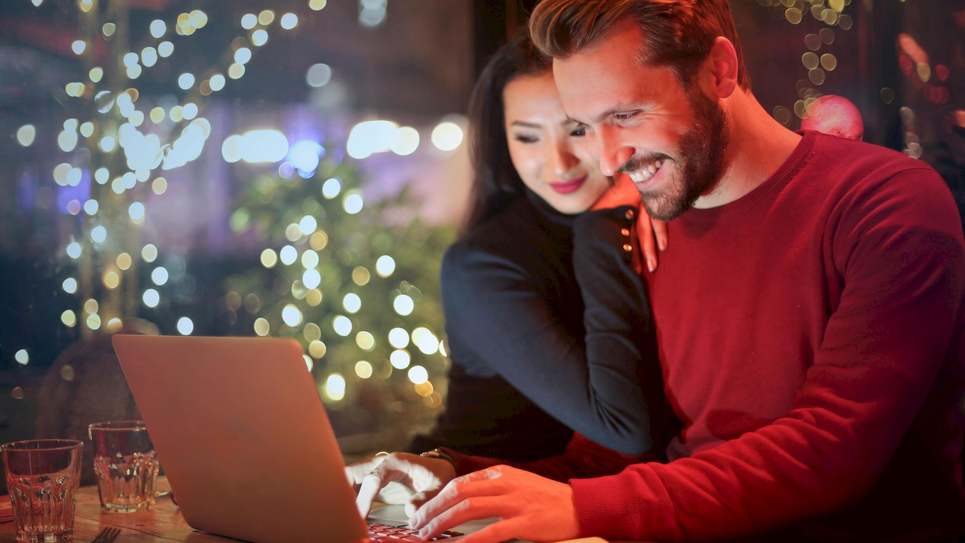A smiling couple is looking at a laptop in a warmly lit cafe with fairy lights and blurred background decorations, appearing happy and engaged.