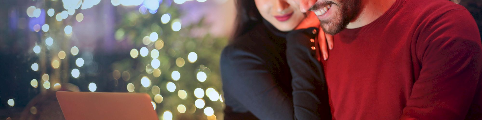 A smiling couple is looking at a laptop in a warmly lit cafe with fairy lights and blurred background decorations, appearing happy and engaged.