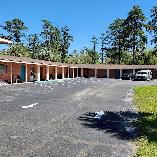 The image shows a single-story motel with a U-shaped layout, a parking lot with two vehicles, and surrounded by trees under a clear blue sky.