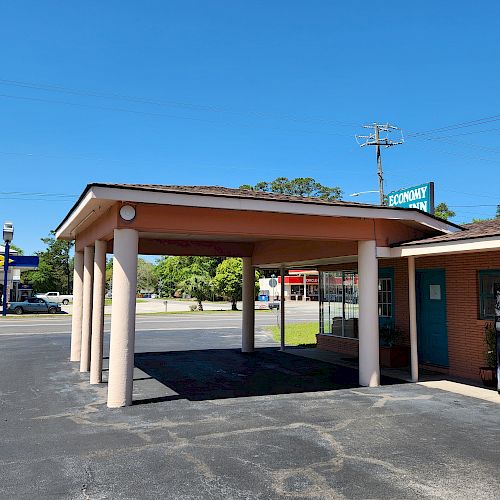 An outdoor area with a covered structure supported by columns next to a brick building, with some signs and a road visible in the background.