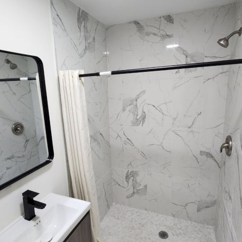 Bathroom with marble-look tiles, shower curtain, mirror with black frame, and sink with black faucet, reflecting the shower.