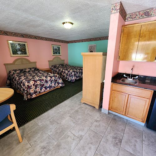 This image shows a motel room with two beds, a small dining table, chairs, kitchenette, microwave, mini fridge, and an armoire, featuring colorful decor.