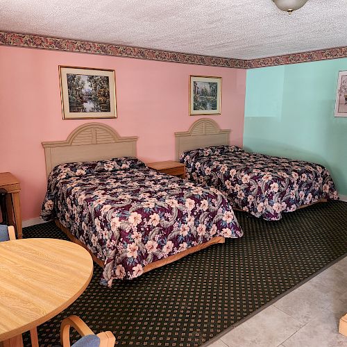 This image shows a hotel room with two beds, floral bedspreads, a small dining table, and wall art. The walls are painted pink and green, ending the sentence.