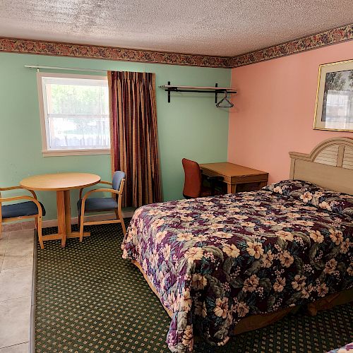 A hotel room with pastel walls, a bed with floral bedding, a table with two chairs, a desk with a chair, and a window with curtains.