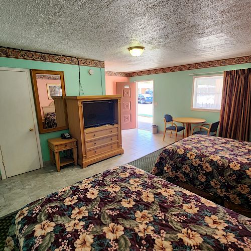 A motel room with two flower-patterned beds, a TV on a cabinet, a table with chairs, and green walls with a door to the outside.