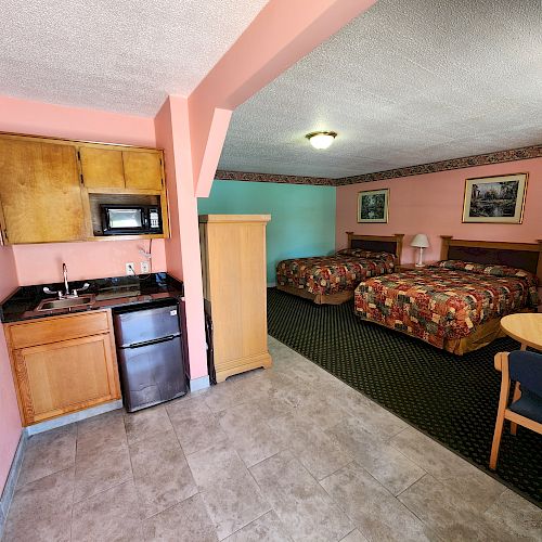 The image shows a hotel room with two beds, a kitchenette with a fridge and microwave, a wardrobe, a table with chairs, and framed pictures on the walls.