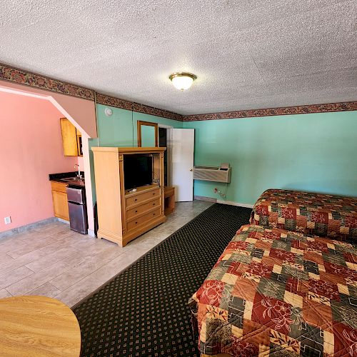 The image shows a motel room with two beds, a TV on a wooden dresser, a small kitchenette area with a microwave and mini-fridge, and multicolored walls.