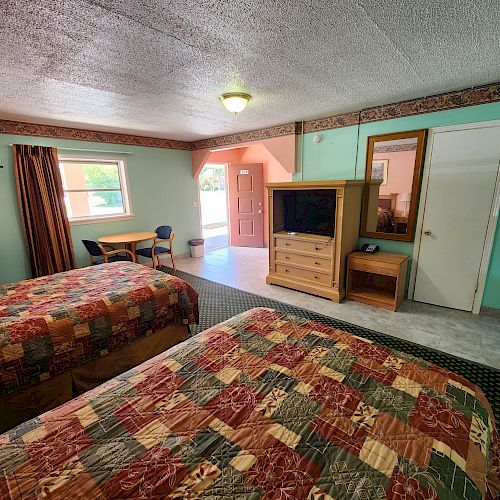 A hotel room with two beds, a TV cabinet, a desk, a window, and a door leading outside. The beds have colorful patchwork quilts ending the sentence.