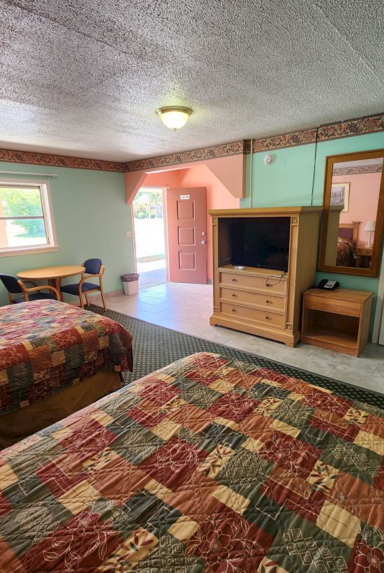 A hotel room with two beds, colorful bedspreads, TV, dresser, desk, chair, and an open door letting in natural light.