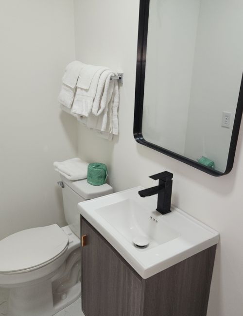 A modern bathroom with a toilet, white towels, and a small sink beneath a mirror. There's green toilet paper on the toilet tank.