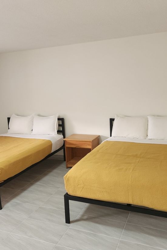 A minimalist room with two beds, yellow bedding, a nightstand, curtains, and hangers on a rail against a plain wall.
