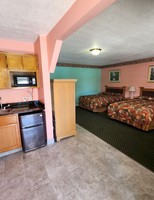 The image shows a hotel room with two beds, a kitchenette area with a microwave and mini-fridge, a small dining table and chairs, and artwork on the walls.